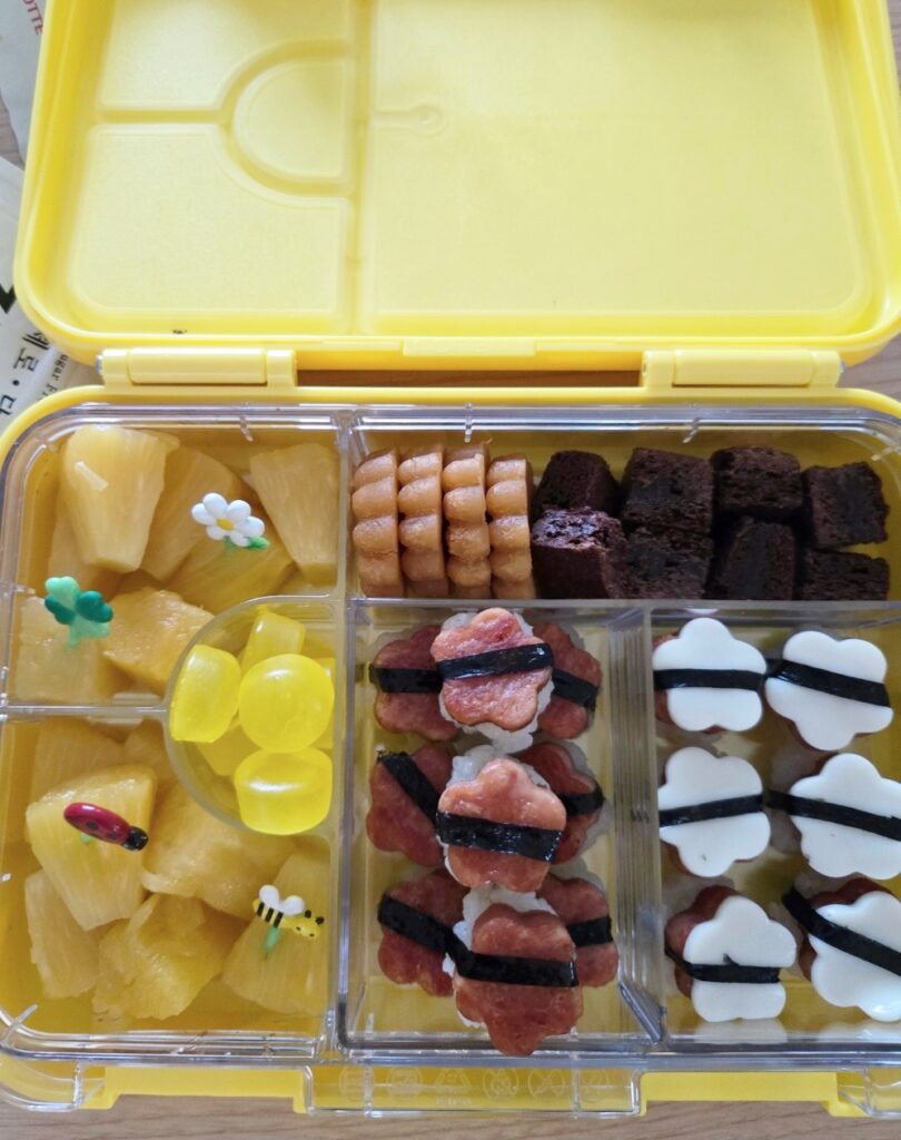 스팸, 치즈 주먹밥, 약과, 브라우니, 젤리, 파인애플이 담긴 리틀그립 야미박스 노란색 도시락 통